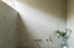 A modern bathroom with white tiled walls and floor, featuring a vase with green leaves and a window letting in natural light.