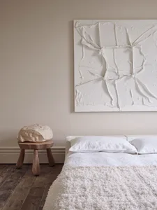 A minimalist bedroom with a textured canvas wall hanging, wooden stool, and white bedding.