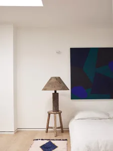 A minimalist bedroom featuring a wooden stool lamp and a white bed.