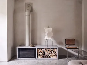 A minimalist living room with a concrete fireplace and wood storage beneath it, featuring a concrete floor and a modern chair