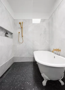 A minimalist bathroom with marble walls and porcelain tiles, featuring a clawfoot bathtub and a walk-in shower with gold fixt