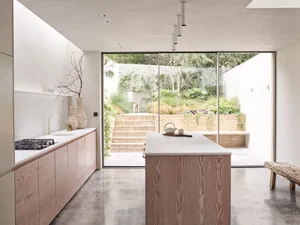 Modern minimalist residential kitchen with concrete island and flooring, large windows overlooking green outdoor space.