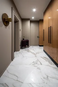 A modern minimalist hallway featuring a sleek marble floor, wooden cabinets, and a wall-mounted light fixture.