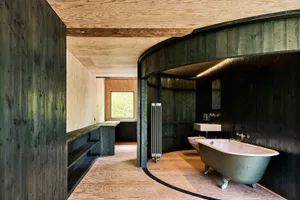 An industrial-style bathroom featuring dark wood walls and floors, with a green bathtub, sink, and radiator, and a window in 