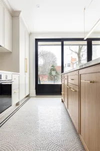 A modern kitchen featuring a tile floor with a wave pattern, wooden cabinets with brass handles, and large windows with a vie