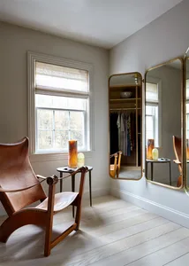 A minimalist living room with natural wood flooring, a leather armchair, a side table with decorative items, and a large mirr