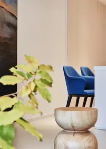 A modern lobby featuring a blue upholstered armchair against a soft pink wall, with a potted plant in the foreground and a wh