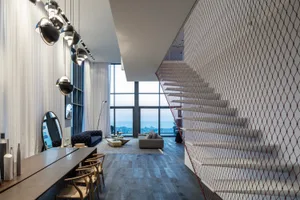 A modern living room with wooden accents, featuring a large table and chairs, a couch, and a staircase with a net railing.
