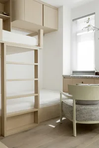 A minimalist bedroom with wooden bunk beds and a chair, featuring a white wall and wooden flooring.