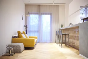 A spacious, modern living room with a yellow sofa, hardwood floors, and sheer curtains.