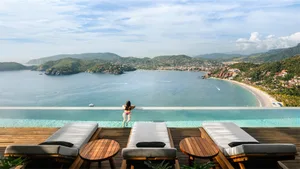 A woman in a swimsuit standing on the edge of a cliffside pool overlooking the ocean and mountains.