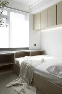 A minimalist white bedroom with a wooden bed, white bedding, and a light-colored cabinet above it, featuring a large window w