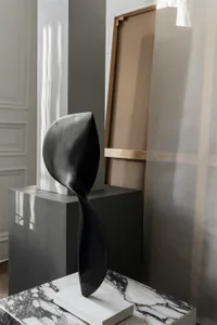 Minimalist black metal sculpture displayed on a white pedestal and marble surface.