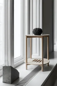 A modern, minimalist side table made of light-colored wood, featuring a round top and a shelf below, with decorative objects 