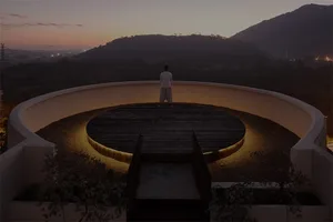 Person standing on a circular wooden platform with illuminated edges, overlooking a mountainous landscape at sunset.