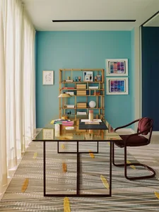 A minimalist home office with a transparent glass and metal desk, a wooden bookshelf, and a maroon chair.