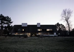 A modern rural house with a thatched roof at night, featuring illuminated windows and a minimalist design.
