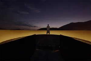 A silhouette of a person standing on a ledge overlooking a calm, golden water body under a twilight sky.