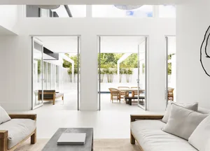 Modern living room with white concrete walls and floor, open doors to an outdoor patio with dining table and chairs, and a mi