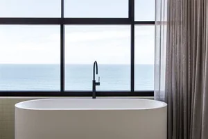A modern bathroom with a white ceramic bathtub, a black faucet, and a large window overlooking the ocean.