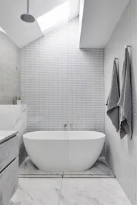 A minimalist bathroom with marble tiles, concrete walls, and a freestanding bathtub under a skylight.