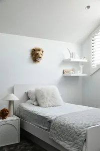 A minimalist bedroom with a white bed, fur pillow, and wooden headboard, featuring a lion's head and a shelf with animal figu