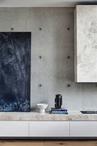 A modern living room featuring a concrete wall with a large dark abstract painting, a white ceramic bowl, a black ceramic vas
