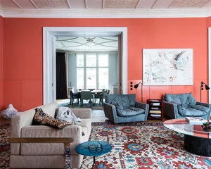 A modern living room with coral-colored walls and a patterned rug, featuring two blue armchairs and a beige sofa.