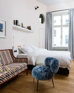 A modern bedroom with a white bed on a wooden floor, featuring a colorful couch and fluffy chair, and a shelf with decorative