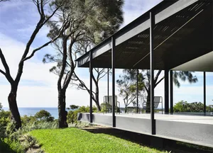 A modern residential exterior with steel beams and a glass wall overlooking the ocean and forest.