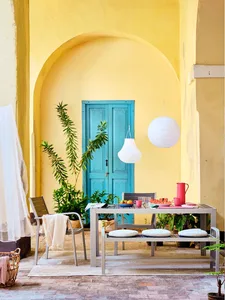 Outdoor dining area with a wooden table, chairs, and hanging lanterns against a yellow and blue arched wall.