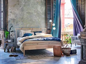 Minimalist bedroom with a wooden bed and blue accents, featuring a rustic wall and large windows with blue curtains.