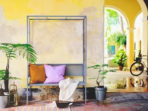 A modern interior decor image featuring a metal frame with purple and mustard pillows on a bench, surrounded by potted plants
