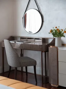 A modern dressing table with a round mirror, a checkered upholstered chair, and a small vase of orange flowers.