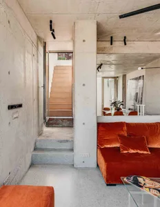 An industrial-style living room with exposed concrete walls and floors, featuring an orange velvet sofa and minimalist decor.