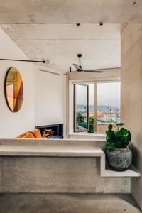 A modern living room with concrete walls, an orange couch, a fireplace, and a large window with a city view.