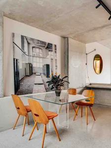 Modern industrial dining room with concrete walls, wooden chairs, and glass table.