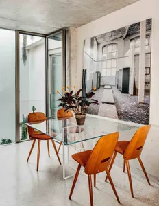 A modern industrial dining room featuring a glass table, wooden chairs, and concrete walls.