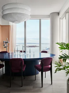 A modern dining room features a round wooden table with purple upholstered chairs, glass candle holders, and large windows ov