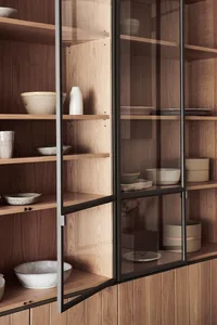 Modern kitchen cabinet with wooden shelves, glass doors, and black metal accents displaying ceramic bowls and plates