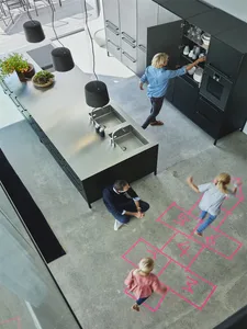 A modern minimalist kitchen featuring a metallic island with glass finishes and a concrete floor, with children playing hopsc