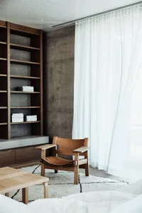 A minimalist bedroom with concrete walls, a leather chair, and a large window with white curtains.