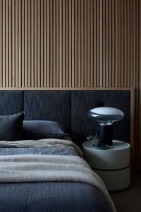 A modern bedroom featuring a bed with a wood-paneled headboard, grey linens, and a bedside table with a lamp and plant.