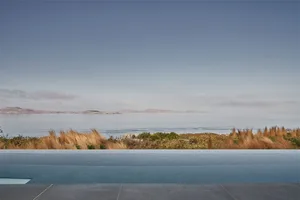A serene ocean view featuring a swimming pool with wind-blown grass in the foreground.