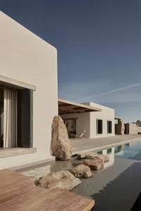 A minimalist residential exterior with a poolside area featuring concrete walls, wooden deck, and stone elements.