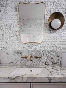 A modern bathroom featuring a marble countertop and sink, with a stylized mural wallpaper on the wall and a brass mirror and