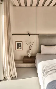 A minimalist bedroom featuring a concrete floor, a grey upholstered bed, a wooden bedside table with a vase of dried flowers,