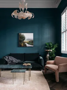 A modern living room featuring a dark green velvet sofa, a marble coffee table, and a pink armchair. The room has dark teal w