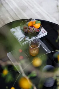 Fruit bowl with grapes, oranges, and leaves atop a stack of magazines on a glass coffee table surrounded by greenery.