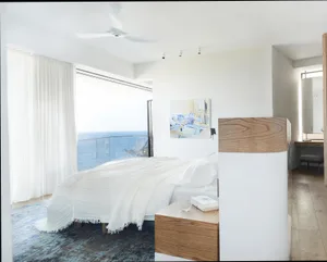 A modern minimalist bedroom featuring white bedding, a wooden nightstand, and a white accent wall with a small abstract paint